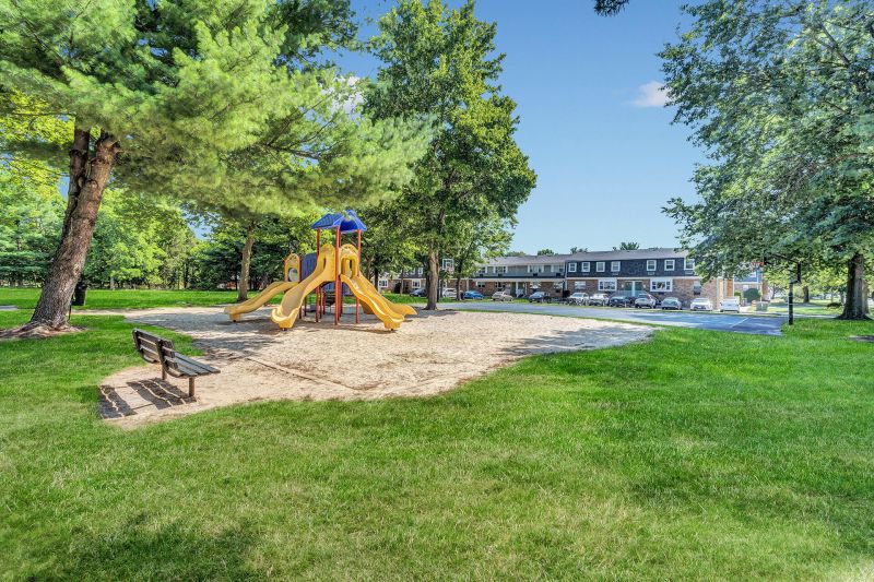Exterior photo of Beekman Garden in Hillsborough, NJ showing the community playground, surrounded by grass and trees with a bench for relaxing