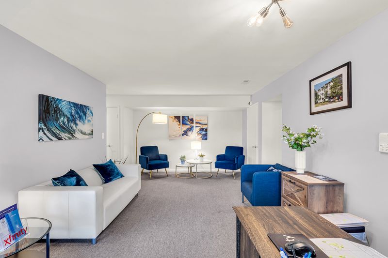 Interior photo of the living room at Green Plaza showing a large four-person couch alongside two accent chairs with side tables. Multiple decorative paintings on freshly painted walls. Grey carpet runs through the entire room.