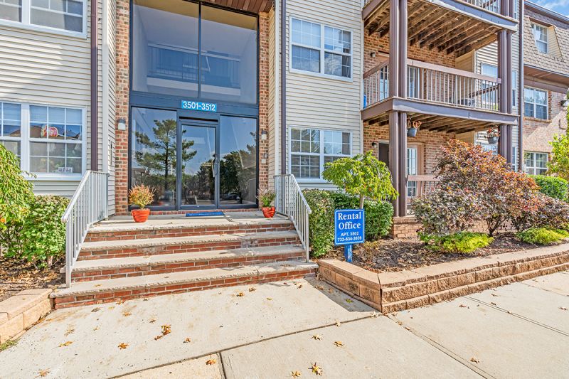 Exterior photo of the rental office entrance at Green Plaza showing exterior finishes including brick, cream siding, and balconies. A brick staircase with white railings leads to the front door of the rental office. Sidewalks and fresh landscaping surround the building.
