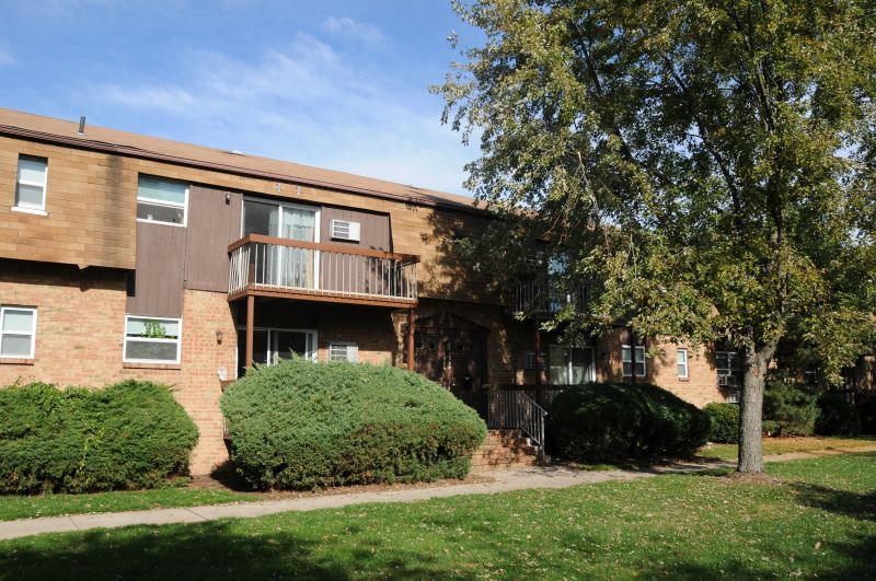 Florida Grove Manor exterior photo shows a two story brick apartment building with both first and second floor balconies. Green grass, bushes and trees landscape the front of the building.