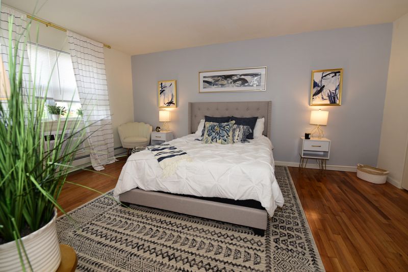 Photo of the primary bedroom with bed, 2 end tables with lamps, a chair and large windows to allow for plenty of natural light.