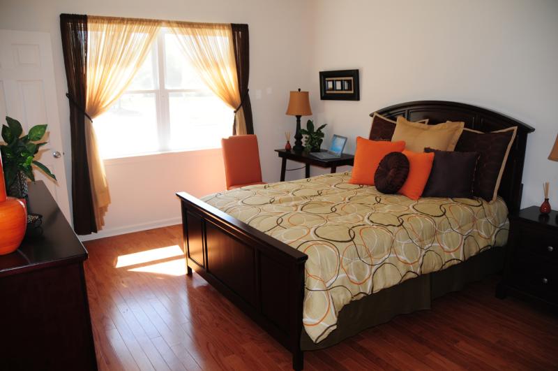 North Brunswick Manor Bedroom photo showing a queen sized bed, a nightstand, a dresser and a small table with a chair. There is a computer on the table and several decorations on the table and dresser. There is a large window to allow natural light to enter the room.