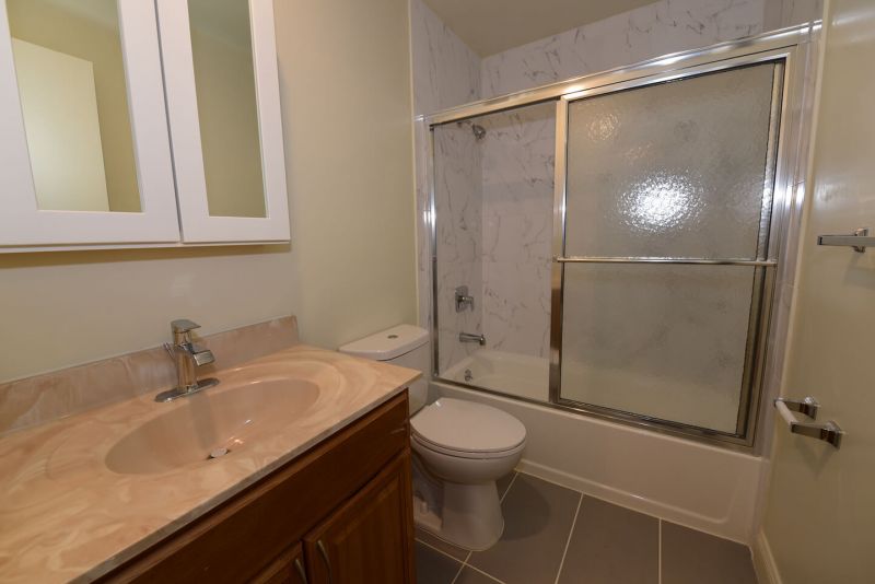 Photo showing a freshly painted renovated bathroom showing new ceramic tile flooring, new tub with shower doors, new toilet, sink, vanity and medicine cabinet.