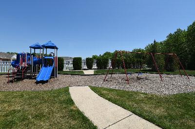 Woodhaven Terrace Playground photo showing a playground and a swing set. The pool can be seen in the background with a white fence around it. Several bushes and trees highlight the landscape.