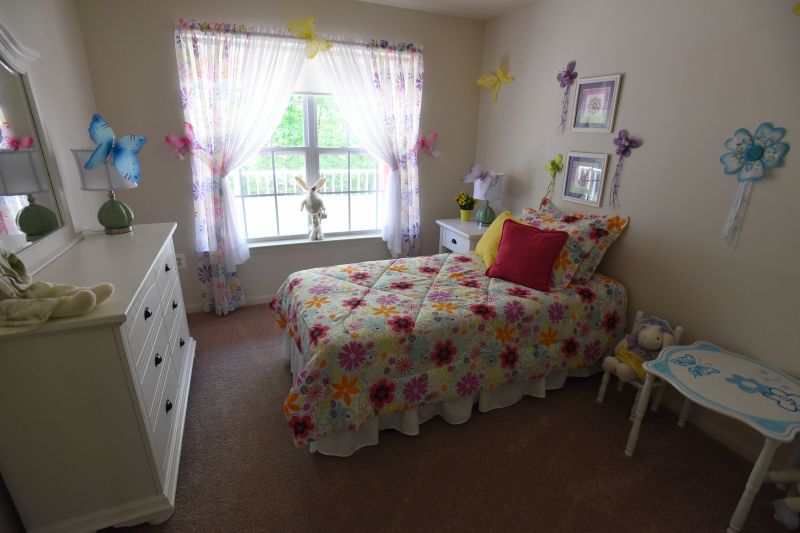 Woodhaven Terrace Bedroom photo showing a bed, a dresser with a mirror, a nightsand and a small table and chair. There is a large window to allow natural light to enter the room. There are decorations and pictures around the room and on the dresser and nightstand.