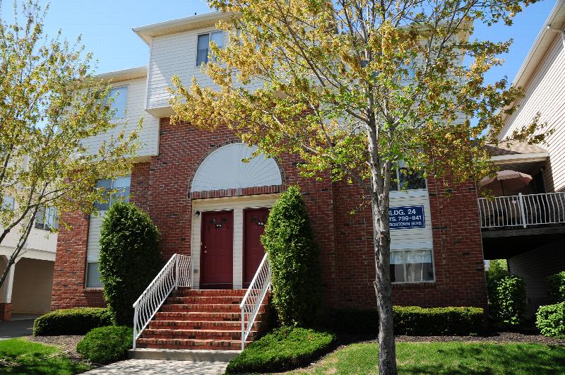Harbortown Terrace exterior photo of an apartment building. There is a stairway leading up to 2 entry doors. The building is red brick with white trim. Trees, bushes and green grass landscape the area.