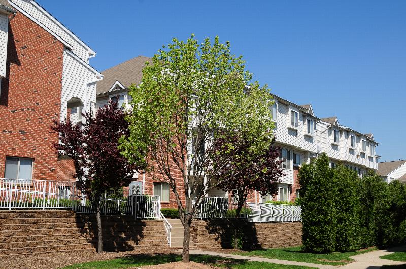 Exterior photo of Harbortown Terrace showing a 3 story apartment building with balconies. There is a sidewalk leading to stairs that bring you up to these buildings. Green grass and mature trees landscape the area.