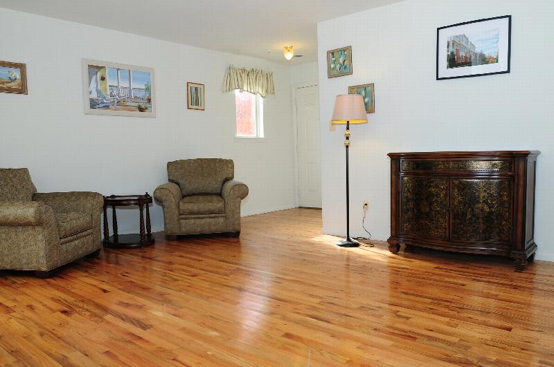 Interior photo of a living room at Harbortown Terrace showing hardwood floors and an entrance door. A seating area, floor lamp, media cabinet and various pieces of wall art decorate the room.