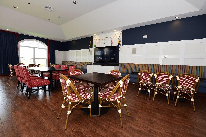Harbortown Clubhouse community room photo showing multiple seating areas and a large screen TV mounted on the wall. There is hardwood floors and a large window to allow for natural light.