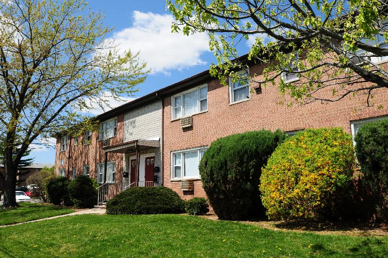 Convery Gardens exterior photo shows a brick apartment building with red front doors and white trim. Green grass, bushes and trees landscape the area in front of the building