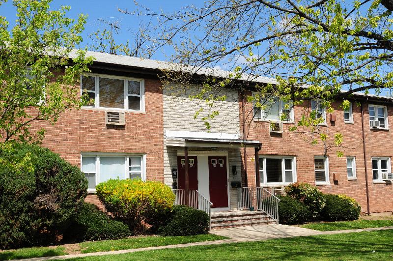 Convery Gardens exterior photo shows a brick apartment building with red front doors and white trim. Green grass, bushes and trees landscape the area in front of the building