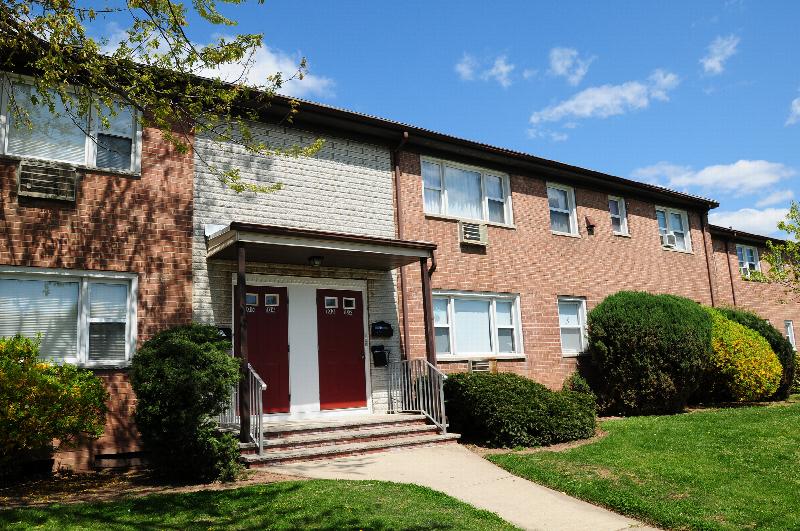 Convery Gardens exterior photo shows a brick apartment building with red front doors and white trim. Green grass, bushes and trees landscape the area in front of the building