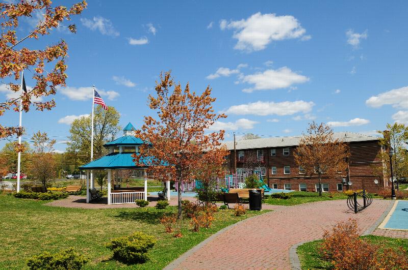Convery Gardens photo shows a nearby gazebo with seating areas. There is a brick paver walkway. There is grass surrounding the area with various trees and bushes.