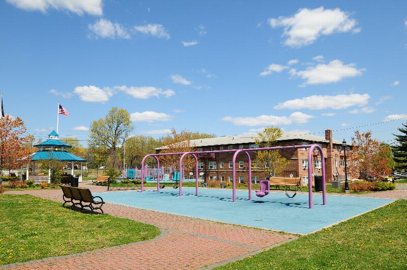 Convery Gardens image of nearby swing sets with benches for seating. A gazebo and apartment building are shown in the distance.