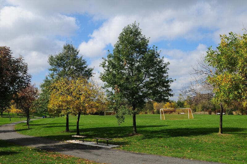 William Warren Park located near Convery Gardens photo of a walking path and benches for resting. Several trees line the path and a soccer field can be seen in the distance.