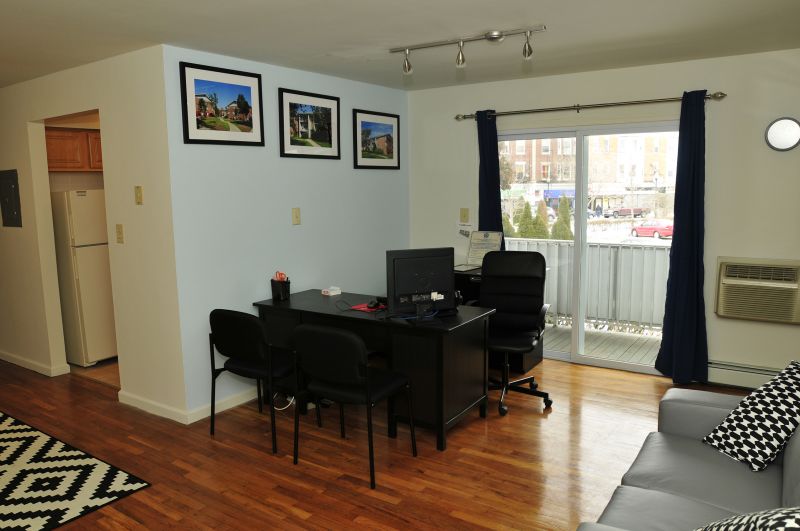 State Fayette Rental Office photo showing a couch, a desk, three chairs and a computer. Hardwood floors run the length of the room. The kitchen can be seen in the background. Sliding glass doors lead to the balcony.