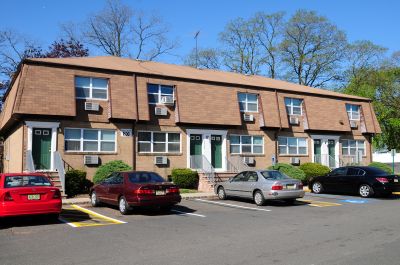 Colonial Gardens exterior photo showing a tan brick apartment building with green doors and white trim. Large green bushes landscape the front of the building. Several cars are shown in the parking lot in front of the building.