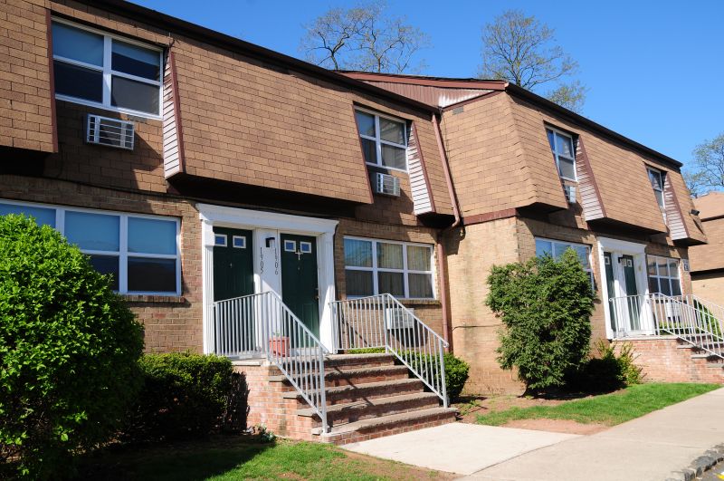 Colonial Gardens exterior photo showing a tan brick apartment building with green doors, white trim and balconies. Several green bushes landscape the front of the building.