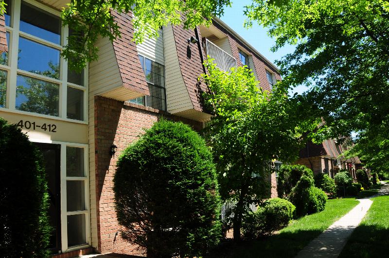Hidden Village exterior showing the apartment building with lush green grass, bushes and trees. A sidewalk leads to additional apartment buildings.