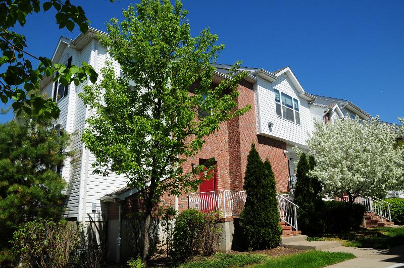 Exterior photo of an apartment building at Harbortown Ports made of red brick and white vinyl siding. Several entry staircases can be seen leading up to red front entry doors. Green grass, bushes and flowering trees landscape the area.