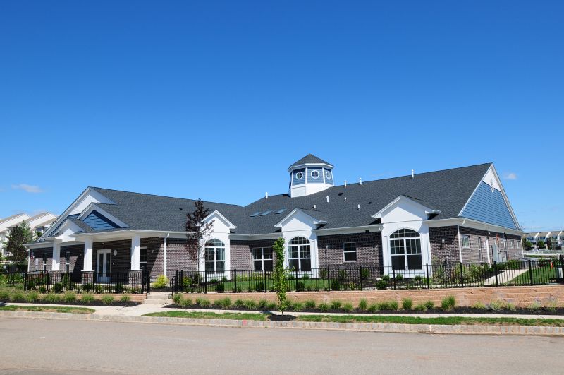Exterior image of the Harbortown Clubhouse showing a large brick building with a covered entrance. There is a black metal fence surrounding the building, and many bushes, plants, flowers and trees landscape the outside.