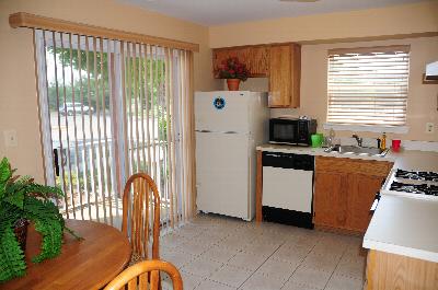 Rivendell Village Kitchen photo showing maple color cabinets, tile flooring and white appliances. There is a round table in with several chairs. There is a large sliding glass door that leads to the balcony. Several decorations are throughout the room.