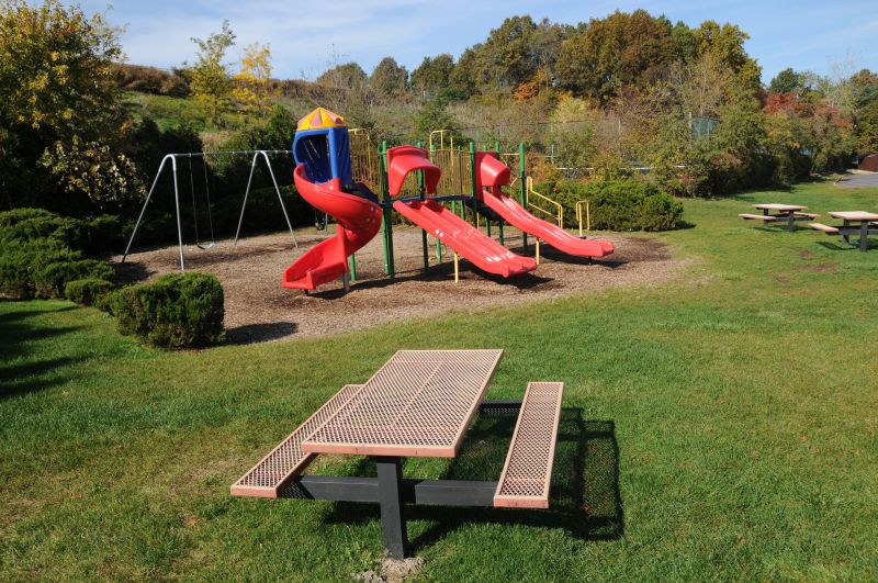 Woodbridge Center Plaza image of the playground area shows swings and a large piece of kids play equipment with several slides. Several picnic table offer seating in the area.
