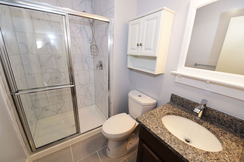 Photo showing a freshly painted renovated bathroom showing new ceramic tile flooring, new tub with shower doors, new toilet, sink, vanity and medicine cabinet.