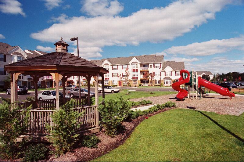 Fox Hill Run exterior image of the community shows a gazebo with childrens play area. The apartment buildings are visible in the distance on the other side of a community parking lot.