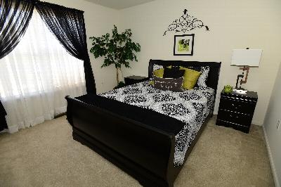 Fox Hill Run photo of a bedroom features a queen sized bed with two end tables and wall to wall carpeting. One of the night stands has a lamp. There are several pieces of wall art on display and a plant in the corner. A large window allows for plenty of natural light.