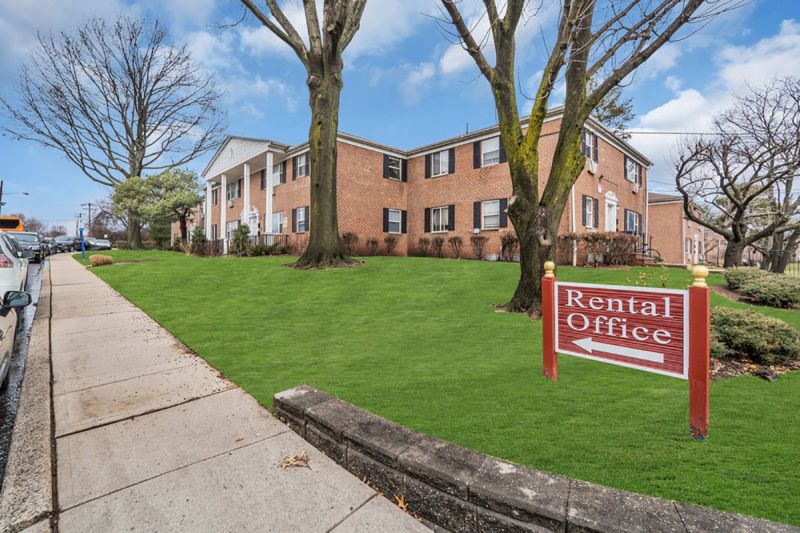 Cloverleaf Gardens photo of the exterior shows a brick building with white trim and red front doors. There is a circular walk way with straight sidewalks leading to each front door. Green Grass and bushes and trees are throughout the area.