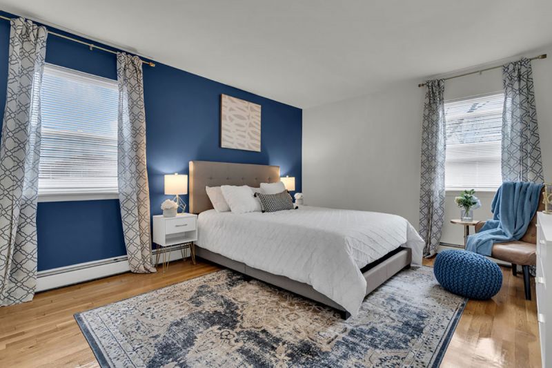 Cloverleaf Gardens photo shows the primary bedroom in a refurbuished apartment with hardwood floors. There is a bed with an area rug underneath, two end tables with lamps and a seating area. Two large windows allow for natural light.