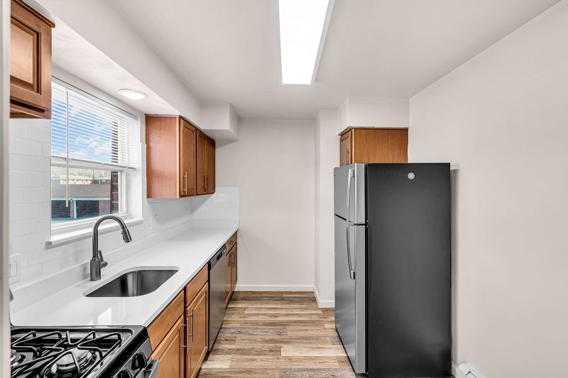 Interior photo of the kitchen at Cloverleaf Gardens showing hardwood floors, brand-new cabinetry, stainless steel appliances, and white quartz countertops complemented by a classic white subway tile backsplash. A large window above the sink fills the space with natural light.