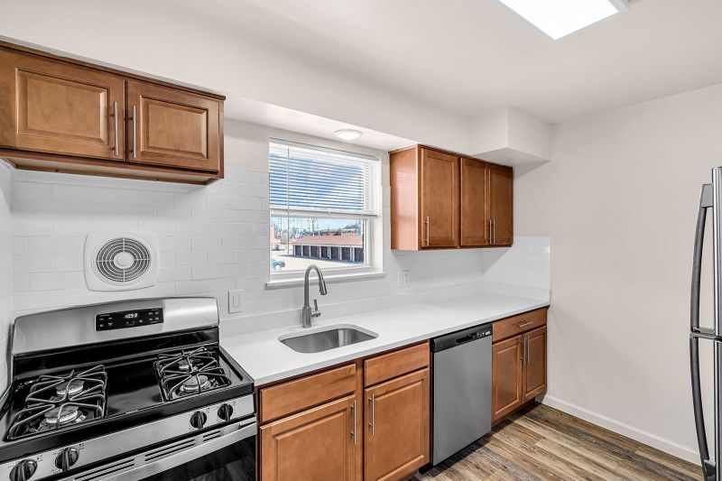 Interior photo of the kitchen at Cloverleaf Gardens showing another angle of the hardwood floors, brand-new cabinetry, stainless steel appliances, and white quartz countertops complemented by a classic white subway tile backsplash. A large window above the sink fills the space with natural light.