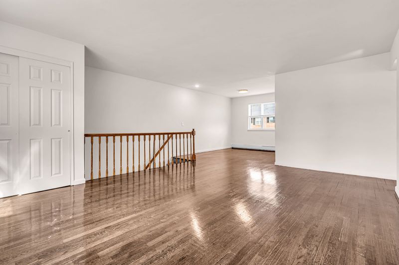 Interior photo of the living room at Cloverleaf Gardens showing the open floor plan connecting the living area to the eat-in kitchen, all featuring luxury hardwood flooring. A staircase leading to the entryway is visible, along with a corner closet with sliding doors. A large window brings in abundant natural light, brightening the space.