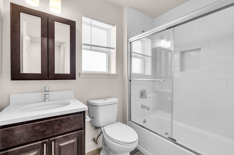 Interior photo of the bathroom at Cloverleaf Gardens featuring luxury hardwood flooring, a bathtub with sleek sliding glass doors, and a dark wood vanity topped with a white quartz countertop and an oval sink fitted with modern Moen Genta faucets. A small window with partially opened blinds above the toilet lets in natural light.
