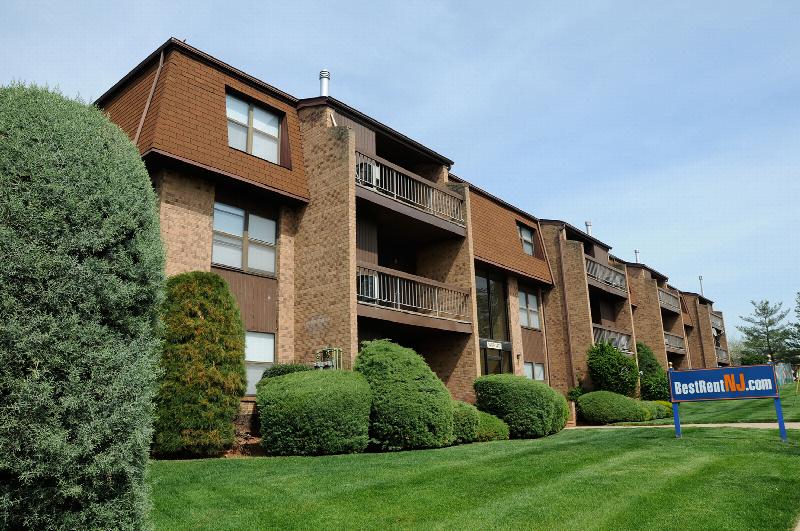Gill Lane exterior photo features a three story apartment building with balconies on each level. A beautiful green lawn and many green bushes landscape the front of the building. A BestRentNJ sign is displayed in the lawn