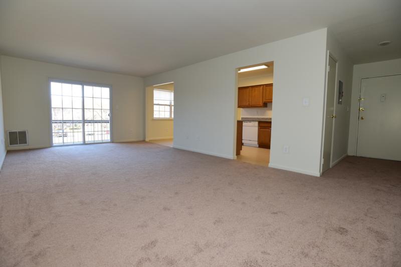 Woodbridge Terrace Living Room photo showing a large room with carpet flooring. Sliding glass doors lead to the balcony and allows plenty of natural light to enter the room. The kitchen and front door can be seen in the background.