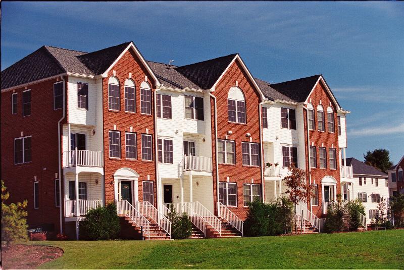 Sun Valley photo of a 3 story apartment building with red brick and white vinyl siding. There are several staircases leading to private entry front doors. There is a large green lawn in front of the building and various bushes and trees.