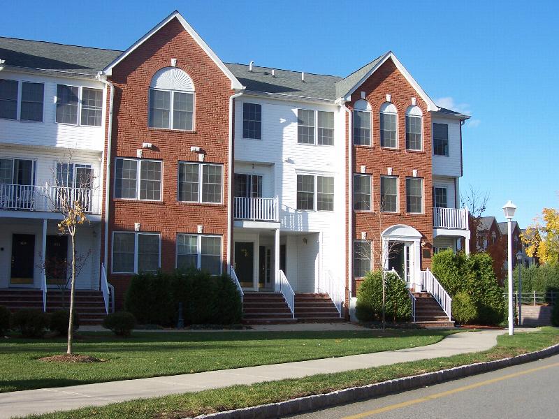 Sun Valley Plaza photo of a 3 story apartment building with red brick and white vinyl siding. There are several staircases leading to private entry front doors. There is a large green lawn in front of the building and various bushes and trees.