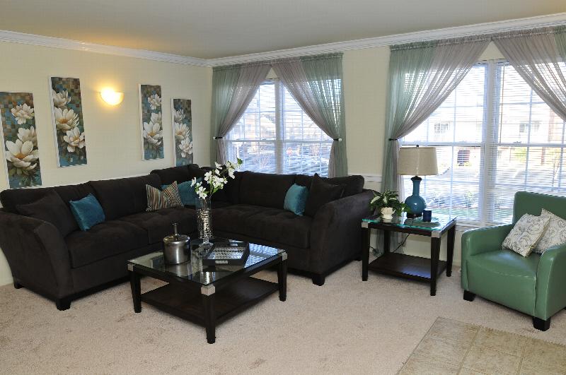 Sun Valley Affordable photo of a living room showing a sectional couch with coffee table, a chair with a side table and various pieces of wall art. There is lush wall-to-wall carpeting and several large windows letting in natural light.