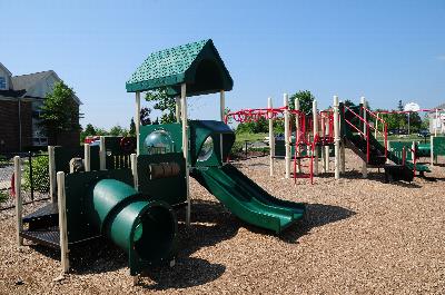 Rivendell Heights Affordable Playground photo showing the playground surrounded by a fence. Several bushes and trees highlight the landscape.