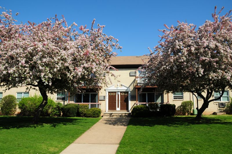 Woodbridge Village exterior show a brick apartment building with brown doors and white trim. A sidewalk leads through the green grass to the front entry. A large pink flowering tree stand tall on both sides of the sidewalk.