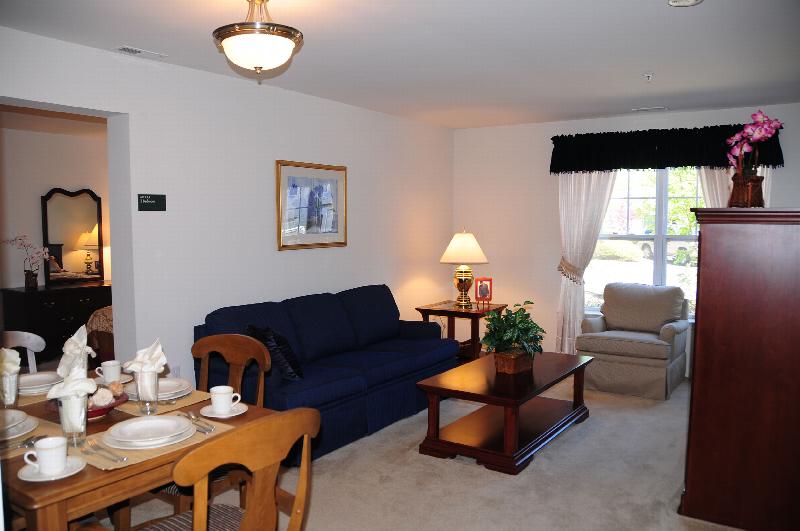 Hyde Park Living Room photo showing a couch with a table and chair. Next to the couch is an end table with a picture and a lamp. There is a dining room table with several chairs and decorations on the table. Carpet flooring runs throughout the apartment. The bedroom can be seen in the background. There is a large window behind the chair to allow natural light to enter the room.