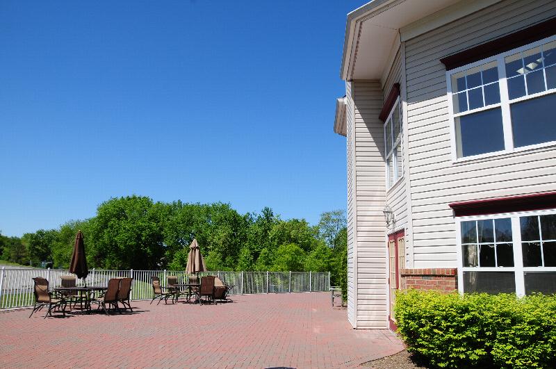 Hyde Park Exterior photo showing the patio with two tables with umbrellas and several chairs. There is a fence around the patio. Several bushes and trees highlight the landscape.