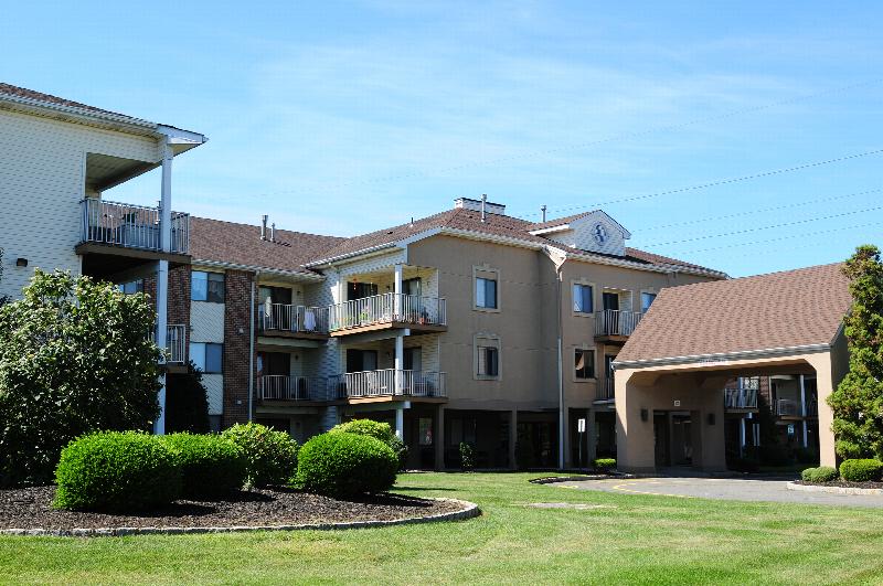 Edison Woods Seniors for ages 55 and over exterior image shows a three story apartment building with a covered entry for cars to drop off, surrounded by green grass, shrubs and landscaping.