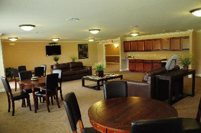 Village at Stratford for ages 55 and above photo of the Community Room shows several couches and tables with chairs as well as a wall mounted TV.