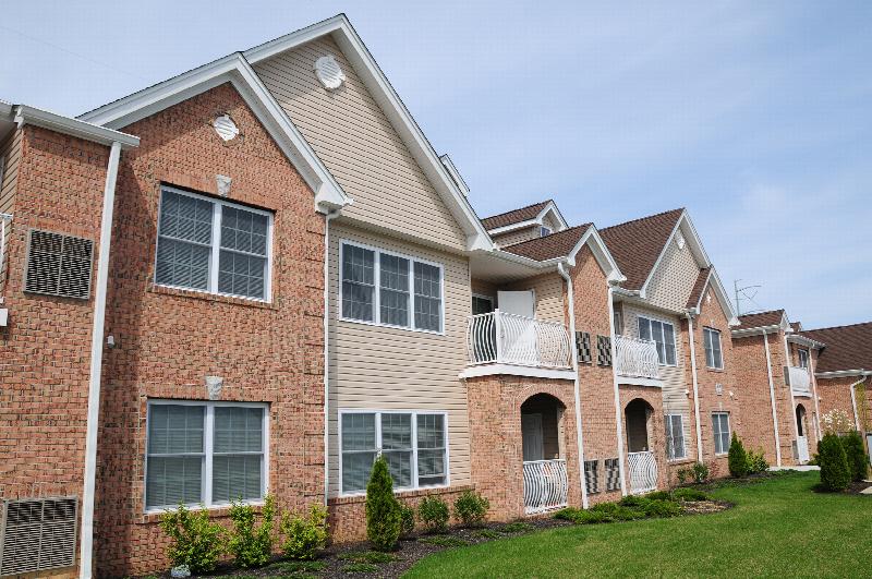 Village at Stratford for ages 55 and above exterior image showing a two story apartment building of reddish brick and cream siding with white trim. Green grass and bushes landscape the area.