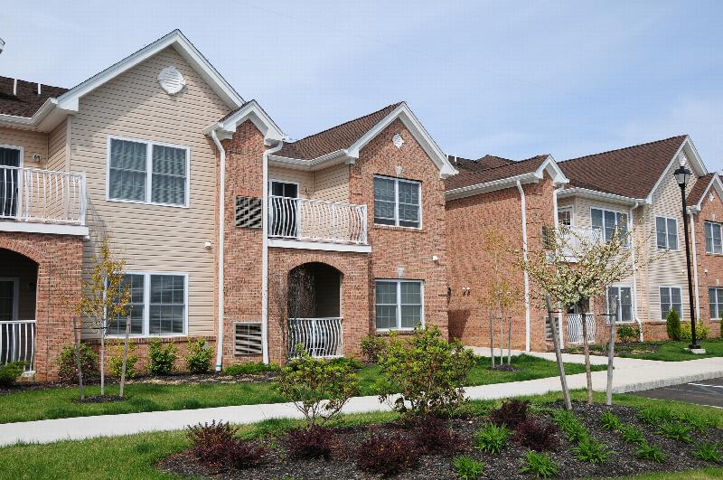 Village at Stratford for ages 55 and above exterior image showing a two story apartment building of reddish brick and cream siding with white trim. Green grass and bushes landscape the area.