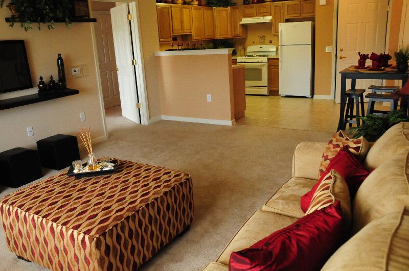 Village at Stratford for ages 55 and above living room photo shows a seating area with a large ottoman and wall mounted TV. The kitchen area can be seen in the background.